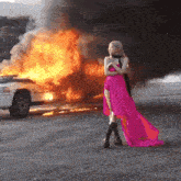 a woman in a pink dress is standing in front of a white car that is on fire