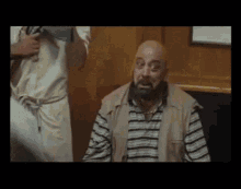 a bald man with a beard is sitting in a room with a man standing behind him .