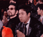 a man wearing sunglasses sits next to another man wearing a black jacket and tie