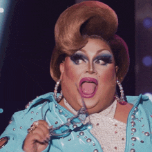 a drag queen is wearing a blue jacket and holding a pair of glasses with her tongue out