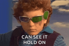 a young boy wearing sunglasses and a vest says can see it hold on .