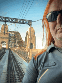 a man with red hair wearing sunglasses stands in front of a bridge