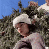a girl wearing a white hat and a sweater is standing in front of a christmas tree .