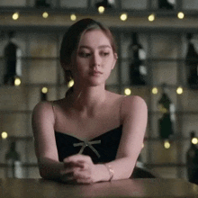 a woman in a black dress is sitting at a table with her hands folded and looking at the camera .