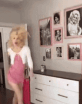 a woman in a pink dress is dancing in a room with a dresser and pictures on the wall