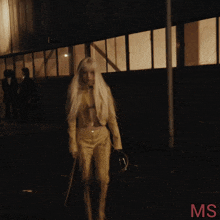 a woman with long white hair is standing in front of a building with the letters ms on the bottom right