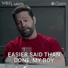 a man in a red shirt is sitting at a table with a glass of wine and says " easier said than done my boy "