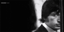 a black and white photo of a man in a suit and tie standing next to a wall .