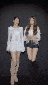 two women are standing next to each other in a dark room . one is wearing a white top and shorts .