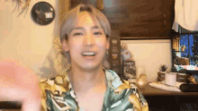 a young man in a hawaiian shirt is waving at the camera while sitting at a desk .