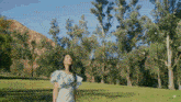 a woman in a white dress stands in a grassy field surrounded by trees