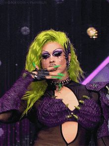 a drag queen with green hair is wearing purple gloves