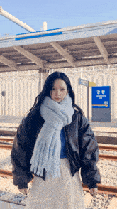 a woman wearing a blue scarf and a black jacket stands in front of a sign that says ganggyeong