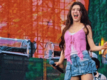 a woman in a pink top and denim skirt sings into a microphone with the name njfaerie below her