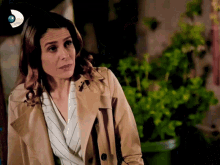 a woman wearing a tan trench coat stands in front of a green potted plant