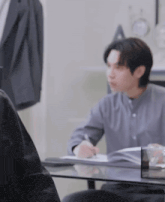 a man is sitting at a table writing on a piece of paper while another man looks on .