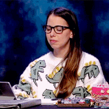 a woman wearing glasses and a sweater with dinosaurs on it sits at a desk