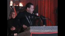 a man stands behind a podium speaking into a microphone while another man stands behind him holding a torch