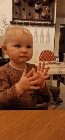 a baby sitting at a table clapping his hands in front of a wine rack