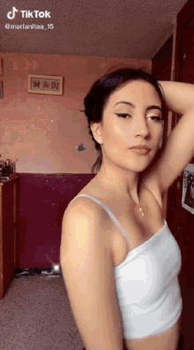 a woman in a white tank top is standing in a room with her hands on her head .