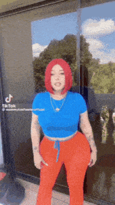 a woman with red hair is standing in front of a window wearing a blue shirt and red pants .