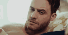 a shirtless man with a beard is laying on a bed looking at something .
