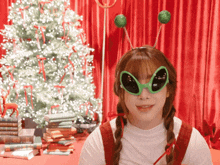 a girl wearing green alien glasses stands in front of a christmas tree and books