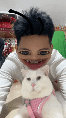 a man with braces on his teeth is holding a white cat in a pink harness
