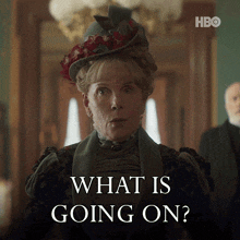 a woman wearing a hat says " what is going on " in front of a man in a suit