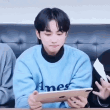 a young man in a blue sweater is sitting at a table holding a tablet .
