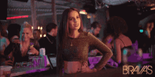 a woman in a crop top stands in a crowded bar with the word bravas on the bottom
