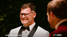 a man in a tuxedo and bow tie smiles while talking to another man in a red jacket