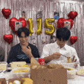 two men are eating noodles in front of a wall with balloons that have the number 115 on it