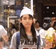 a girl wearing a hat and a t-shirt with the word pacific on it is standing in a crowd of people .