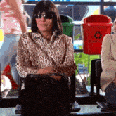 a woman wearing sunglasses and a leopard print shirt is sitting in a chair in front of a recycling bin .