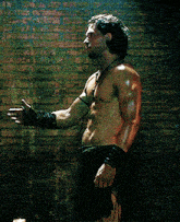 a shirtless man stands in front of a brick wall with his arm outstretched