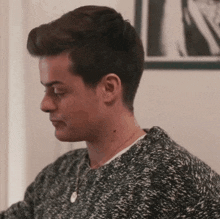 a young man wearing a sweater and a necklace is sitting in front of a wall .