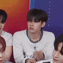a group of young men are sitting in front of a colorful wall . one of the men is wearing a cross necklace .