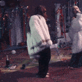 a woman in a fur coat sings into a microphone in a messy room