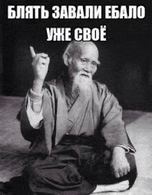a black and white photo of a man with a beard sitting on the floor giving a middle finger .
