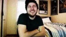 a man with a beard is sitting in a bedroom with his arms crossed .