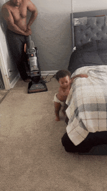 a shirtless man is standing next to a vacuum cleaner and a baby