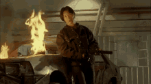 a man is holding a gun in front of a car that is on fire