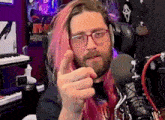 a man with long pink hair and glasses is pointing at the camera while sitting in front of a microphone .