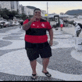 a man in a red and black shirt and black shorts is standing on a sidewalk .