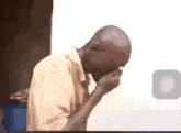 a bald man is covering his face with his hand while sitting in front of a white wall .