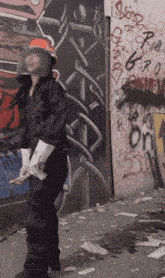 a woman wearing a hard hat and gloves is standing in front of a wall with graffiti on it including the word r.