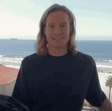 a man with long hair is standing in front of a window overlooking the ocean .