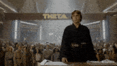 a man stands in front of a crowd under a sign that says theta