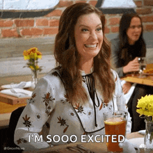 a woman is smiling while sitting at a table with a glass of beer and says i 'm sooo excited .
